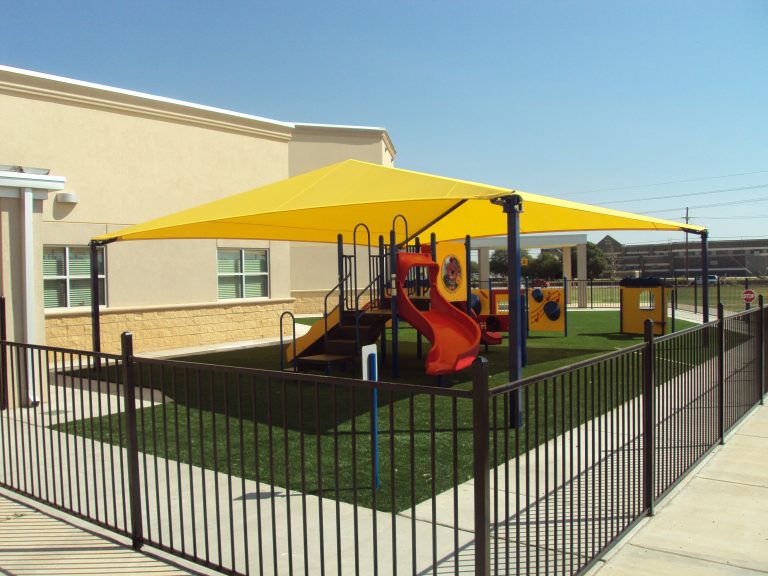 Yellow hip roof shade structure with four black posts covering playground equipment including a red slide and climbing panels