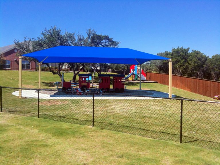 Rectangular hip roof shade structure with blue fabric and four beige posts covering outdoor playground equipment