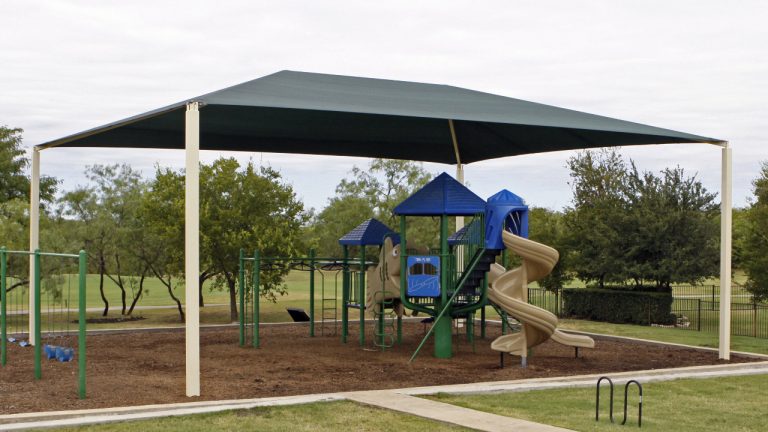 Rectangular hip roof shade structure with four posts covering playground equipment, surrounded by trees and grass