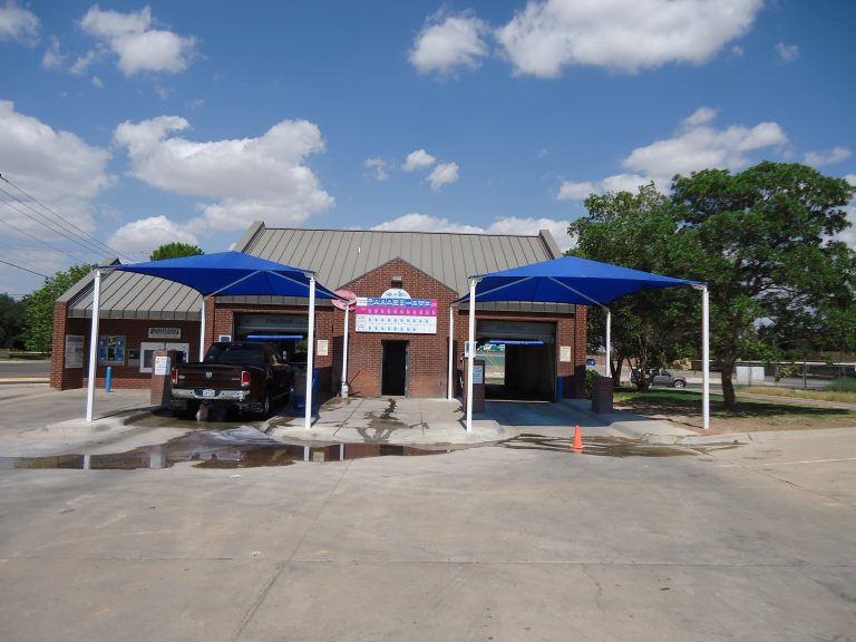Two blue hip roof shade structures with four white posts each covering car wash bays beside a brick building under a partly cloudy sky
