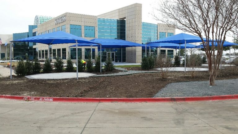 Four-post hip roof shade structures with blue fabric tops over concrete pads in an outdoor commercial setting