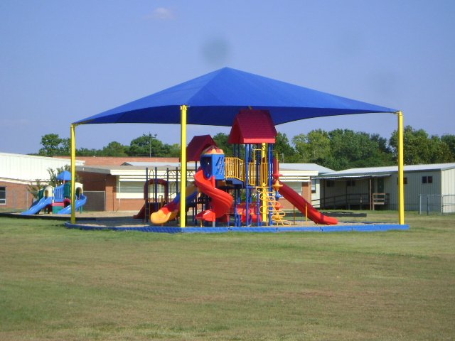Blue pyramid roof shade structure with four yellow posts covering colorful playground equipment on grass field