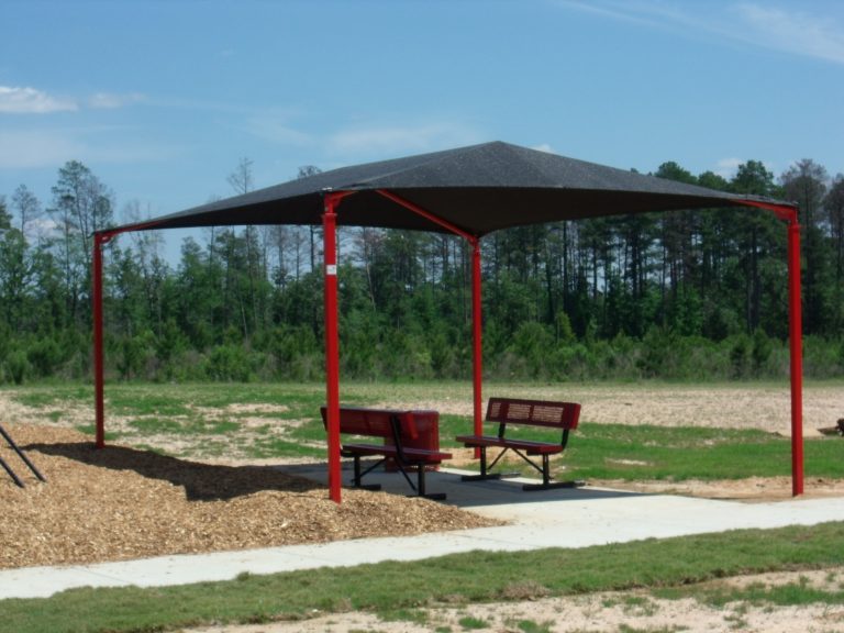 Rectangular hip roof shade structure with red steel posts and black fabric canopy over park benches in outdoor setting