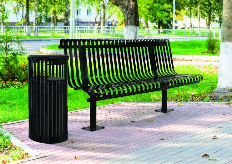 Black metal outdoor bench with rolled vertical slats and backrest beside matching trash receptacle on paved park path