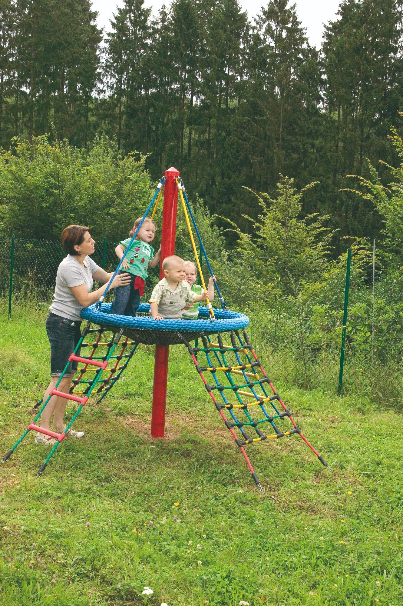 Colorful rope climbing structure with circular nest platform, children playing and adult assisting outdoors on playground equipment