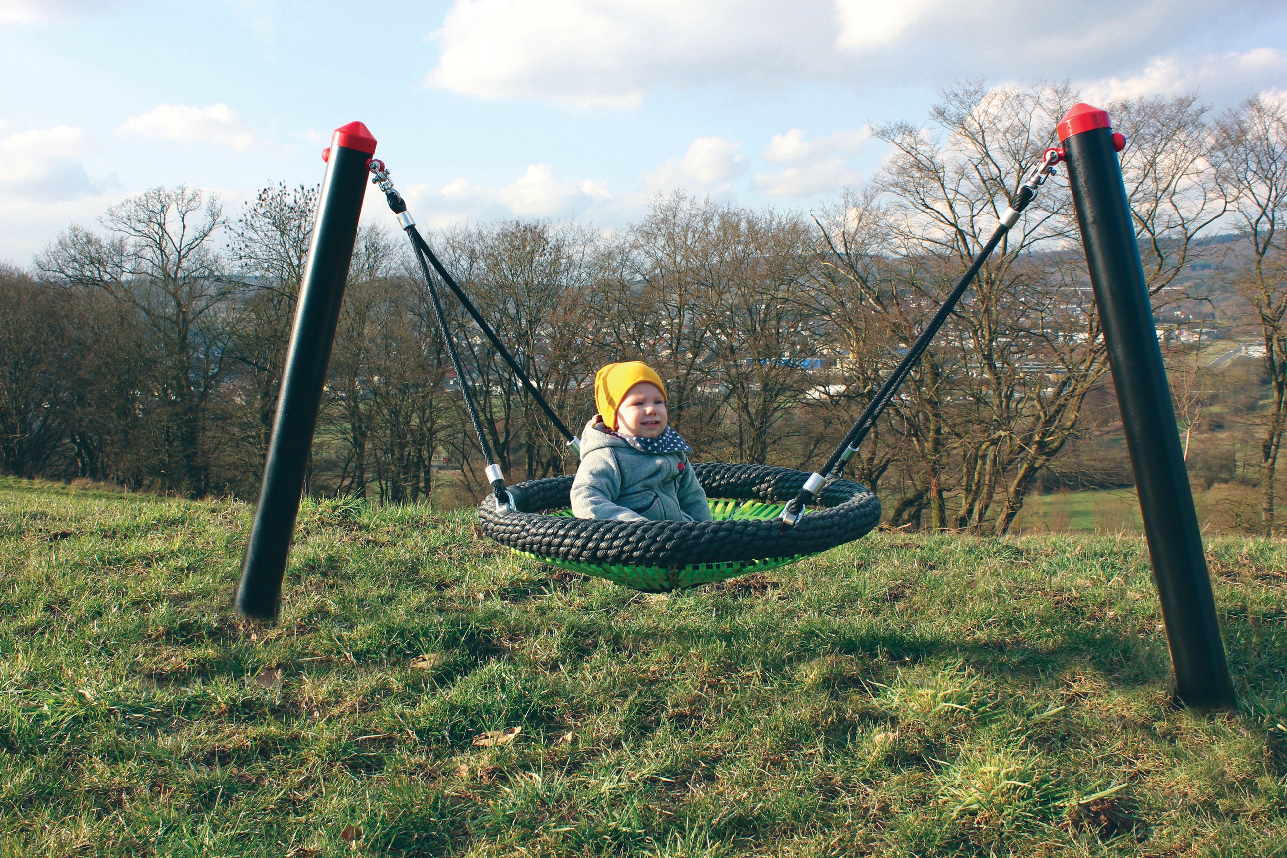 Child sitting in green and black bird's nest mini cradle swing with black posts on grassy hill under blue sky