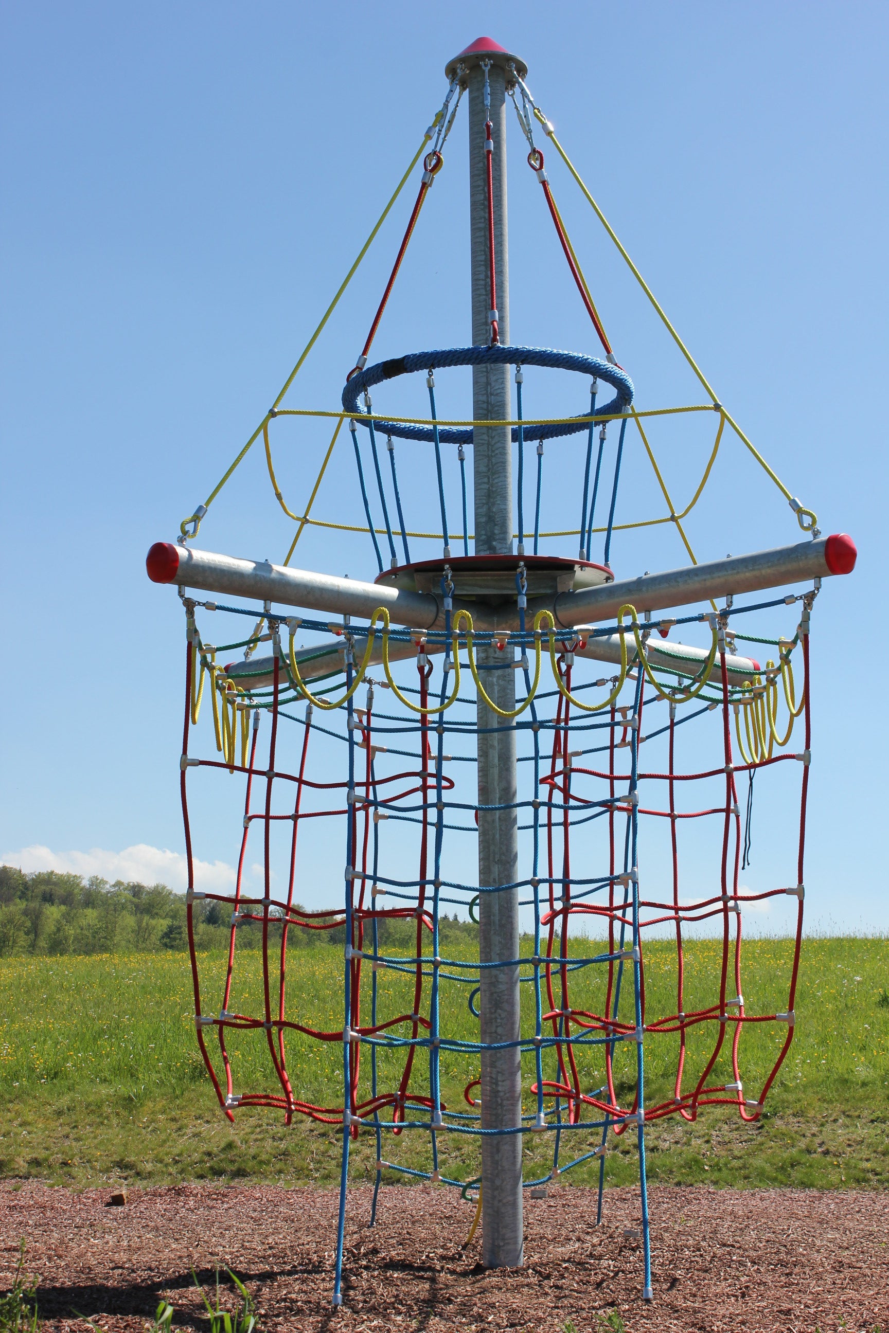 Colorful rope climbing tower with red, blue, and yellow nets and metal frame on playground mulch under clear sky