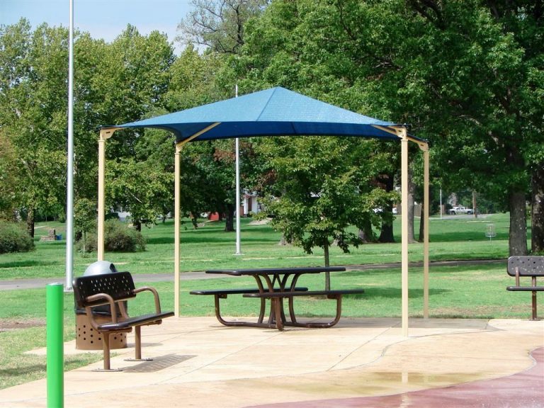 Blue pyramid-shaped fabric shade with four beige posts over picnic table in green park setting