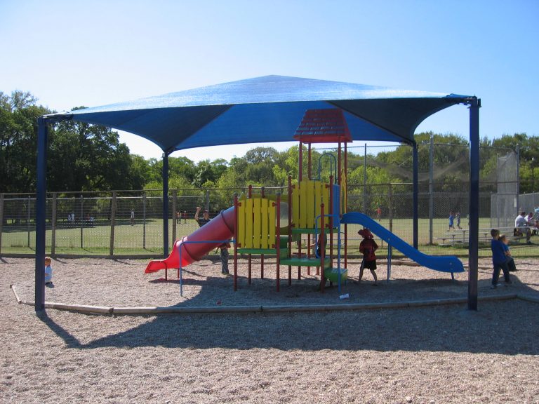 Blue pyramid fabric shade structure with four black posts covering colorful playground equipment and children playing beneath.