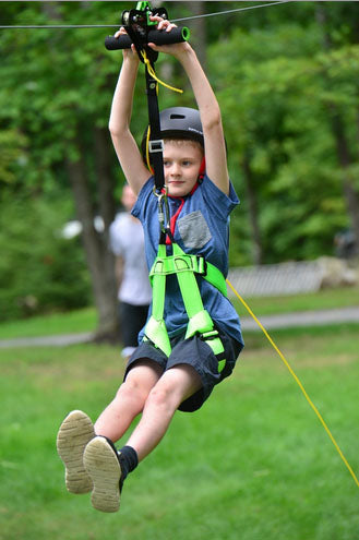 Child wearing helmet and green harness on Alien Flier X3 Zip Line Base Kit trolley in grassy outdoor park setting