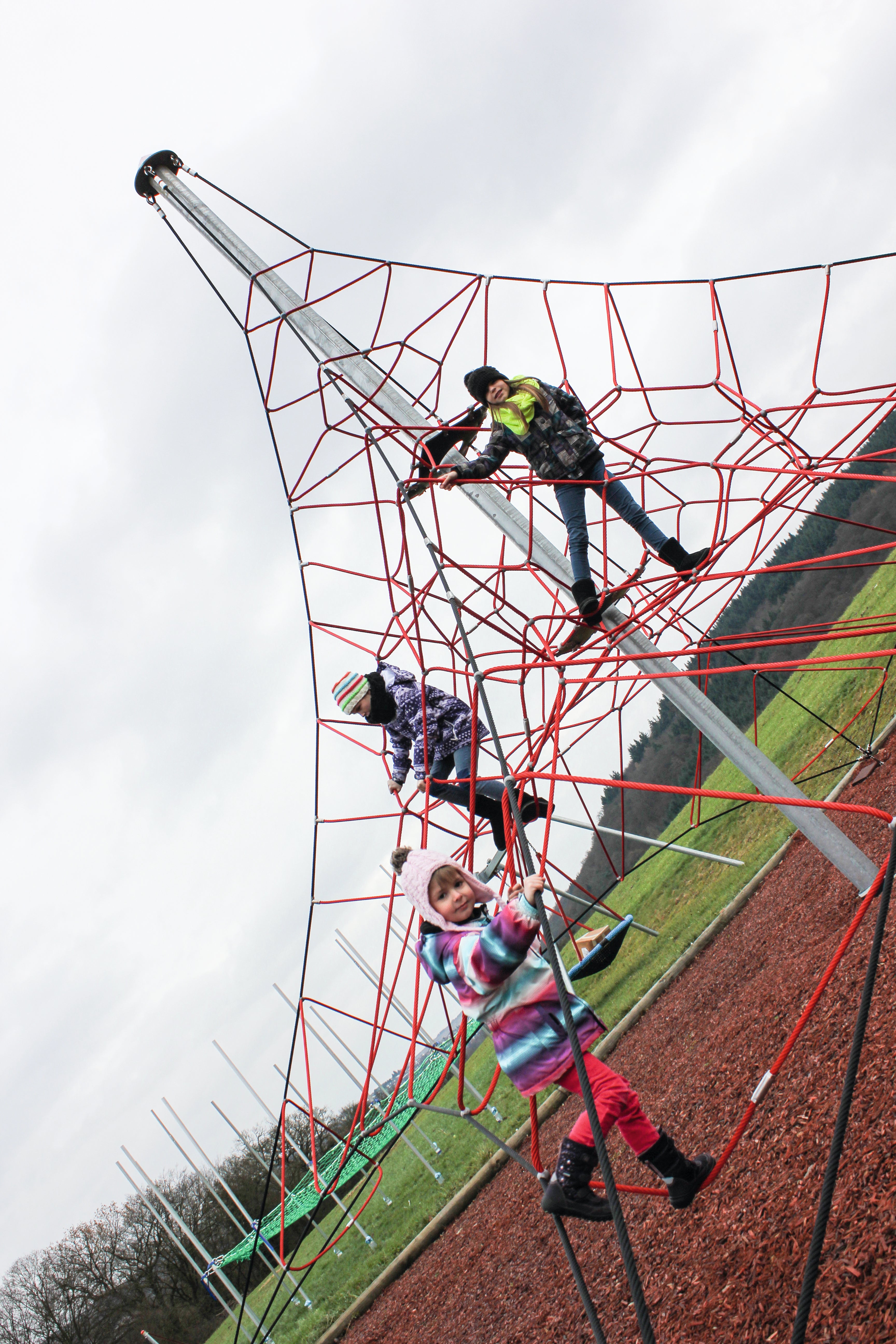 Children climbing a tall red rope net playground structure with metal support poles on a cloudy day