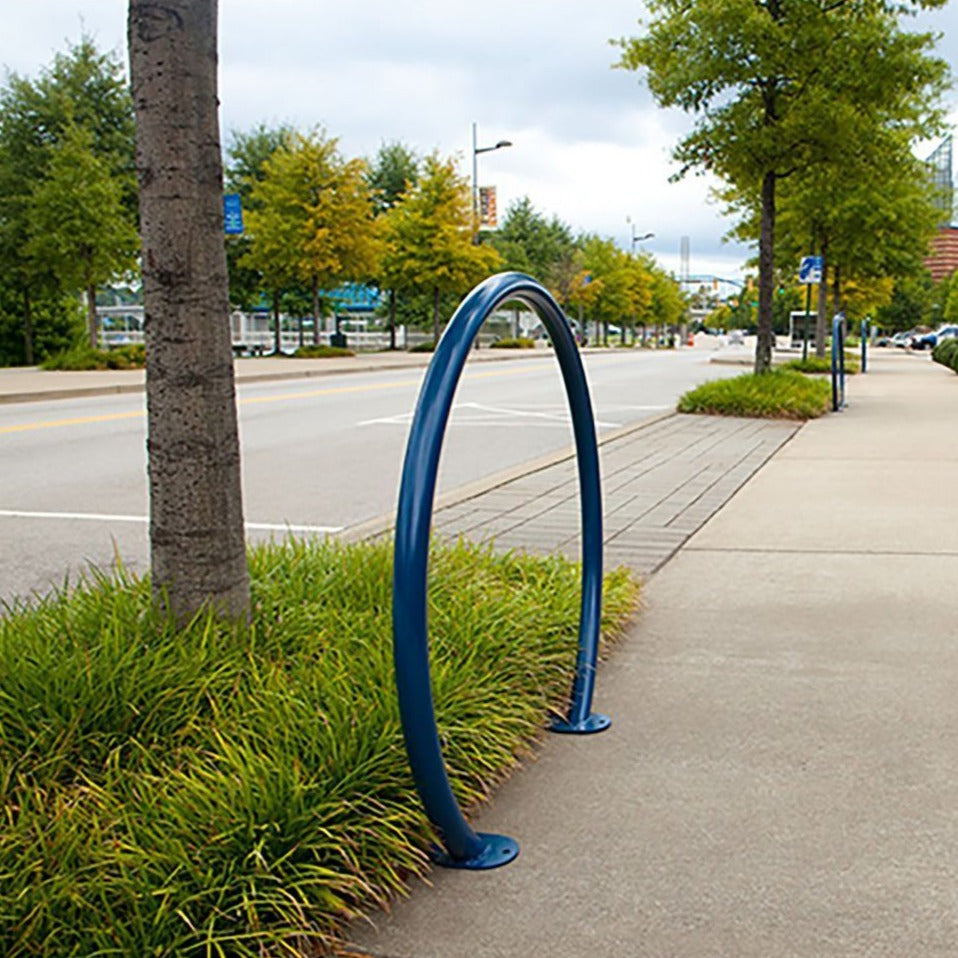 Blue large round tube bike rack installed on sidewalk next to landscaped greenery and street trees