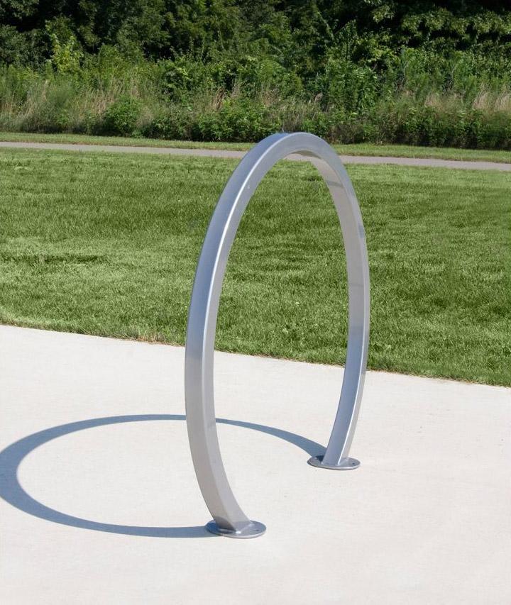 Modern silver metal bike rack with large hoop design mounted on concrete near grass and trees