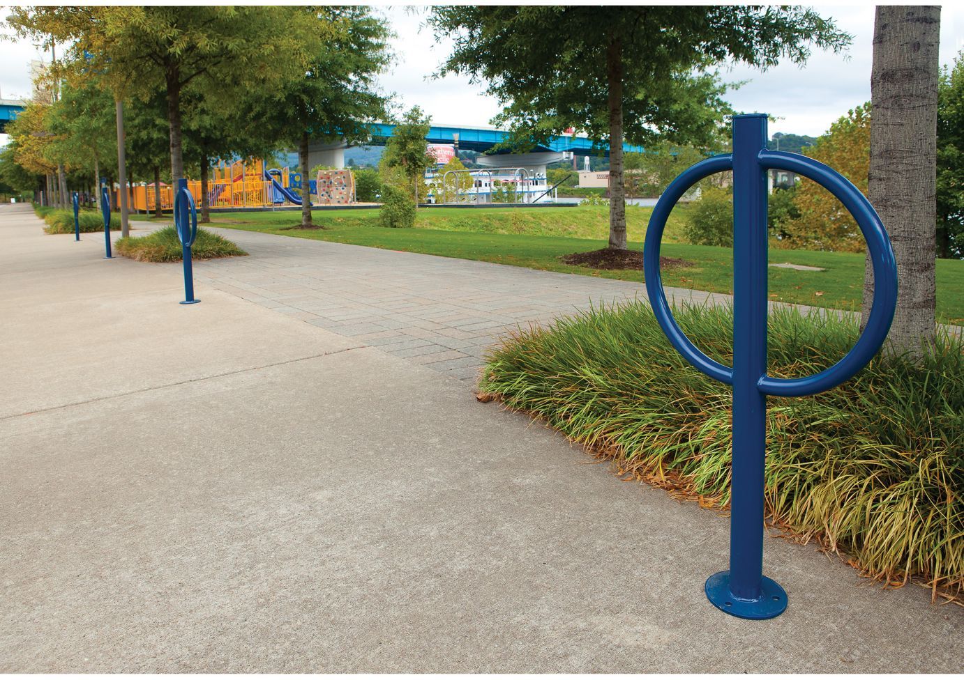 Blue steel bollard-style bike rack with circular ring along paved park pathway near trees and playground
