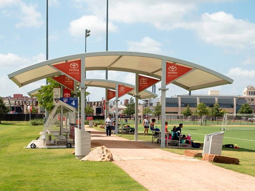 Cantilever shade structure with arched roof covering sports sideline benches and seated people