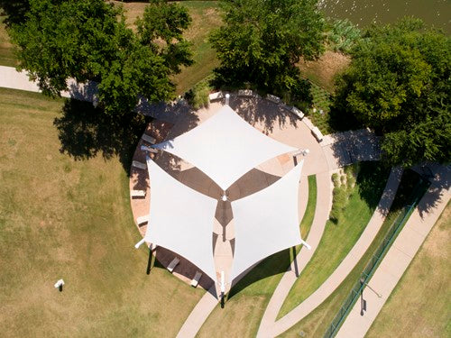 Overhead view of a white 4-point hypar sail shade with four columns casting shadows on circular paved area surrounded by grass and trees