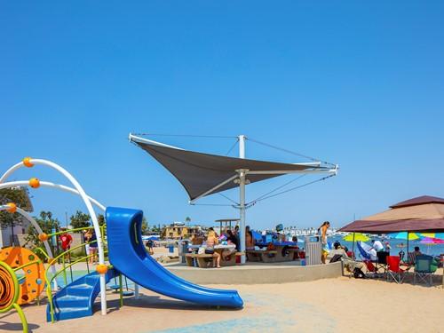 Aurora Single Post Shade Structure with tensioned fabric top shading picnic area near playground and beach setting