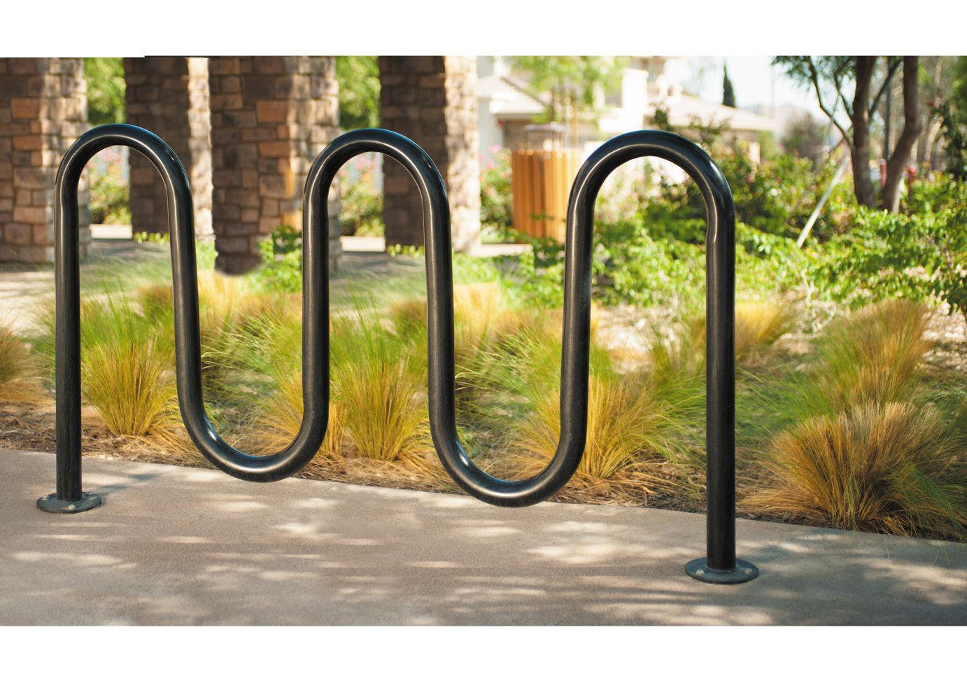Black tubular steel bike rack with five loops mounted on concrete near stone pillars and landscaped greenery
