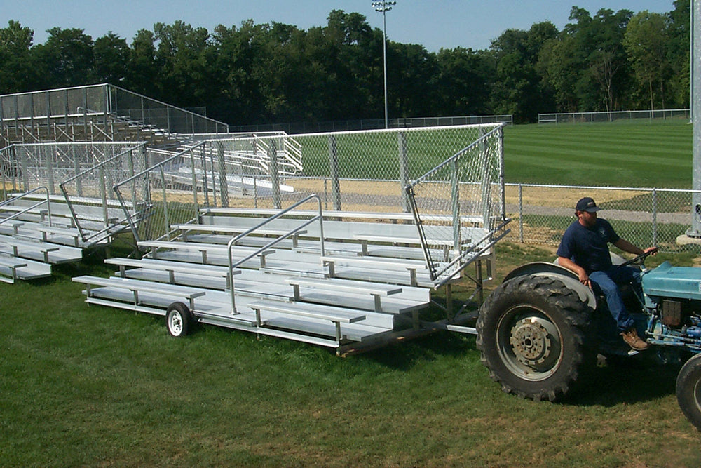 Five-row portable aluminum bleachers with chain-link guardrails, handrail aisle, and transport wheels on grass field