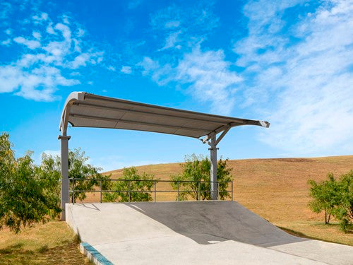 Cantilever shade structure with arched roof over concrete skate ramp in outdoor park setting