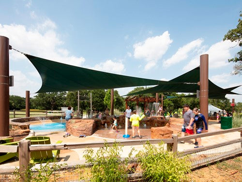 Four-point hypar sail shade with dark fabric stretched over tall columns providing angled shade at outdoor splash pad