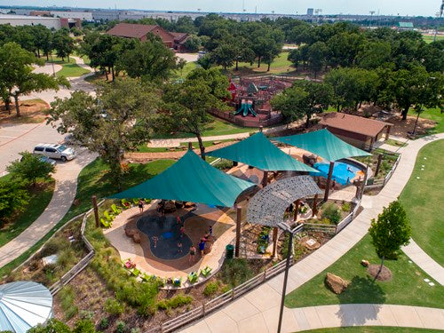 Four teal hyperbolic sail shade structures over a playground splash pad with curved walkways and surrounding greenery