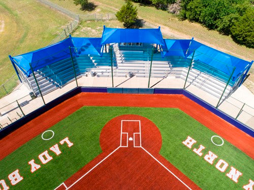 Overhead view of blue cantilever hip roof shade structures covering bleachers at softball field with green turf and red clay infield