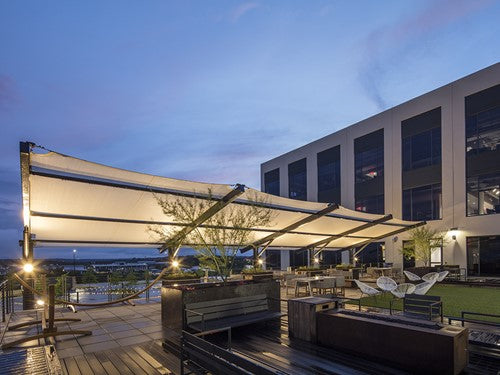 Slanted cantilever wing cabled shade structure with tilted fabric top and two grey columns over outdoor seating at dusk