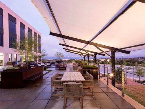 Outdoor patio with cantilever wing cabled shade structure, wicker chairs, wooden table, modern building facade