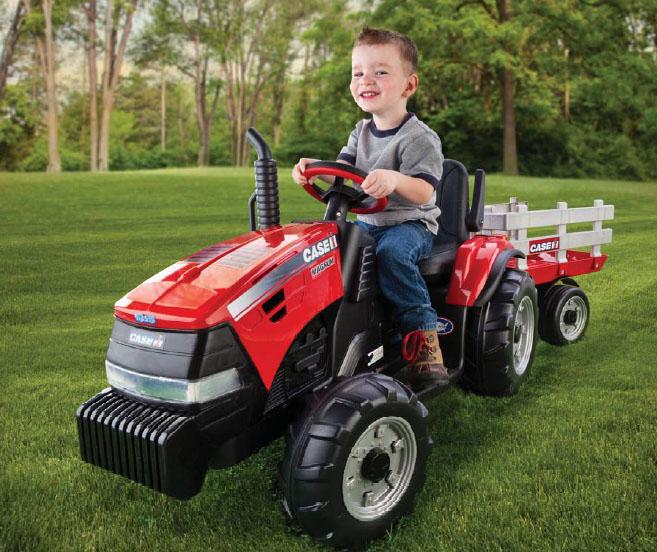 Child riding red 12-volt ride-on tractor with large detachable stake-side trailer on grass