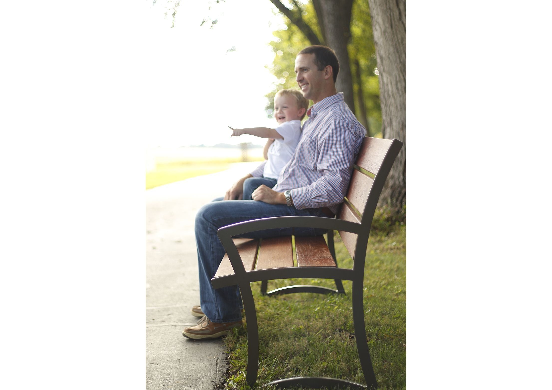 Outdoor Ipe wood bench with steel frame, occupied by a man and child, set on grass beside a paved path under trees