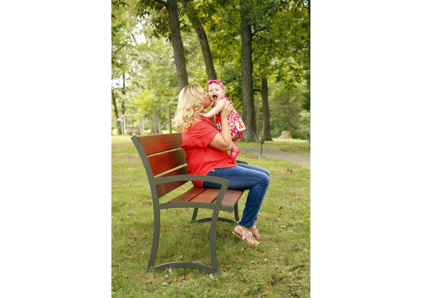 Outdoor park bench with dark metal frame and polished Ipe wood slats, woman in red holding smiling baby in floral dress nearby