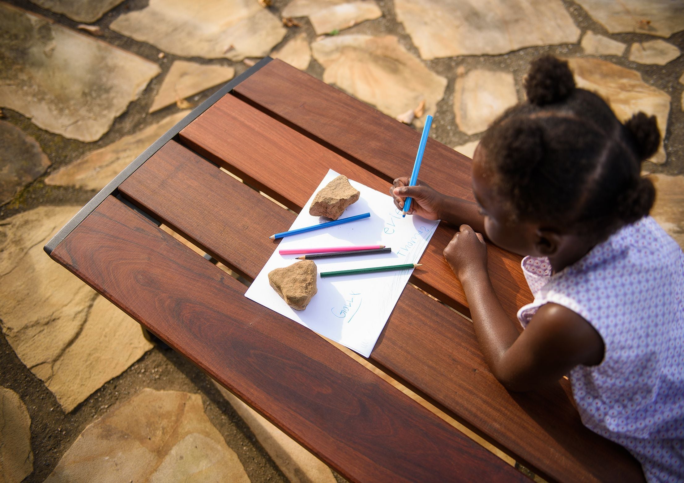 Overhead view of a child drawing with colored pencils on a wooden Ipe wood bench with steel end frames on stone pavement