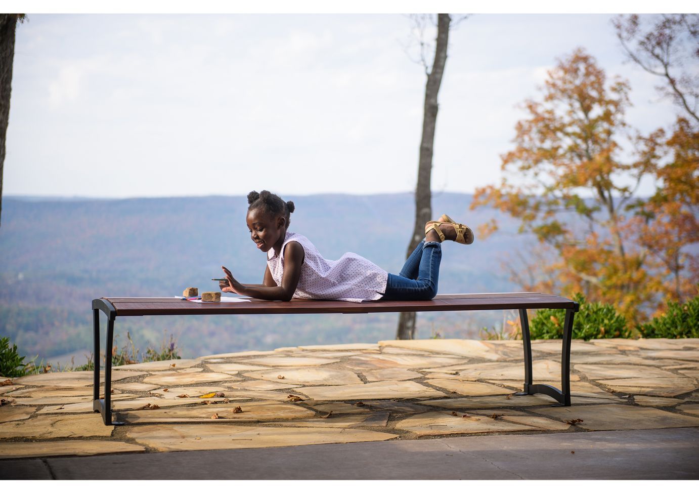 Modern powder-coated steel bench without back on stone patio with child lying on it and autumn trees nearby