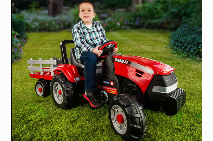 Child on red pedal tractor with large black wheels and detachable stake-side trailer on green grass