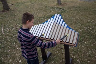 Child playing on blue-trimmed outdoor metallophone mounted on black stand in grassy play area