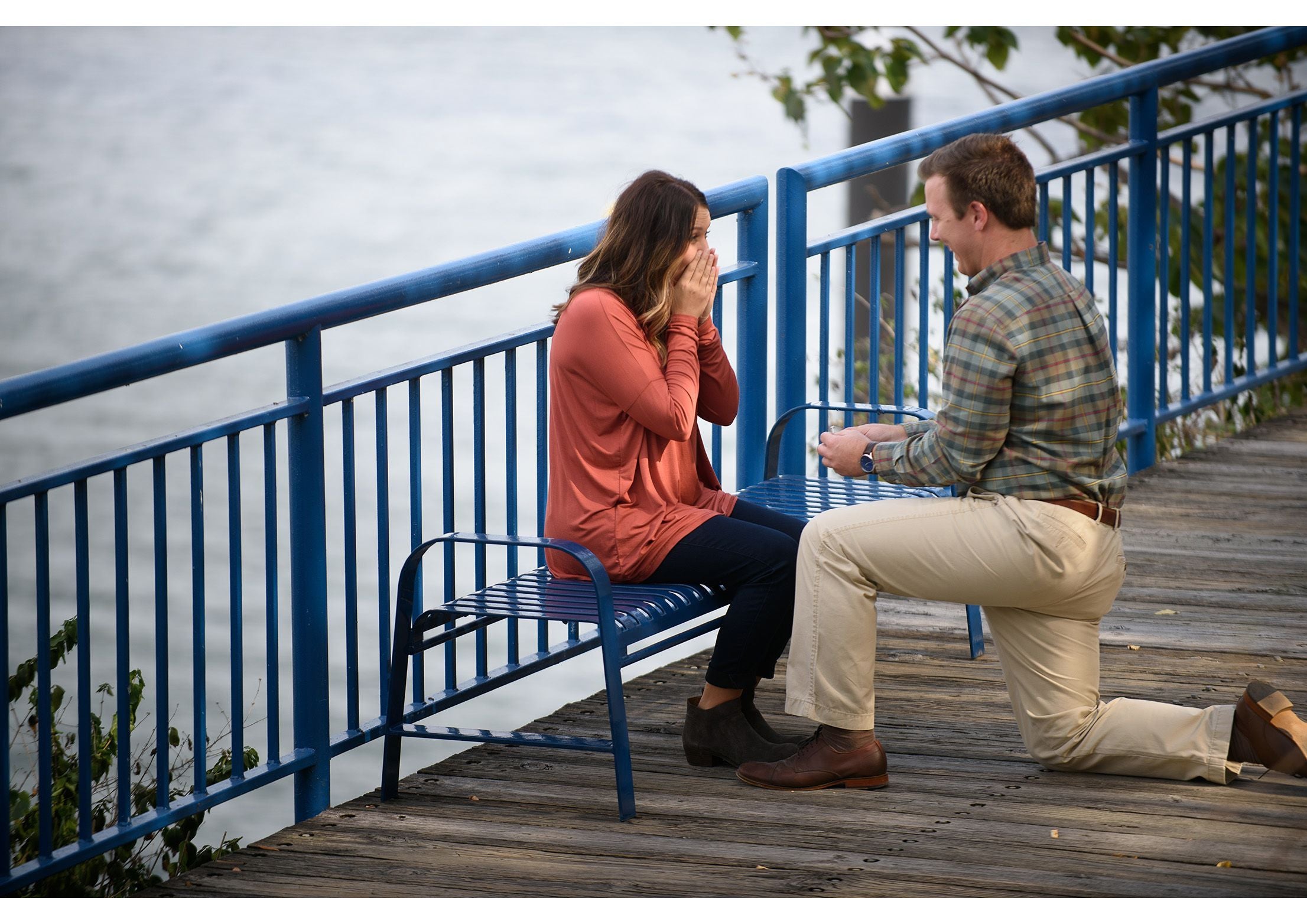 Blue metal backless outdoor bench on wooden dock by water, with a kneeling man and seated woman reacting emotionally