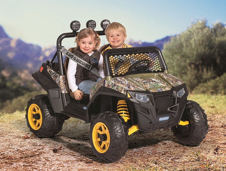 Two children seated in a camo-patterned off-road ride-on vehicle with yellow wheels on a dirt path