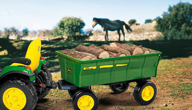 Green John Deere toy farm wagon loaded with logs, attached to a riding tractor on grass with horses nearby