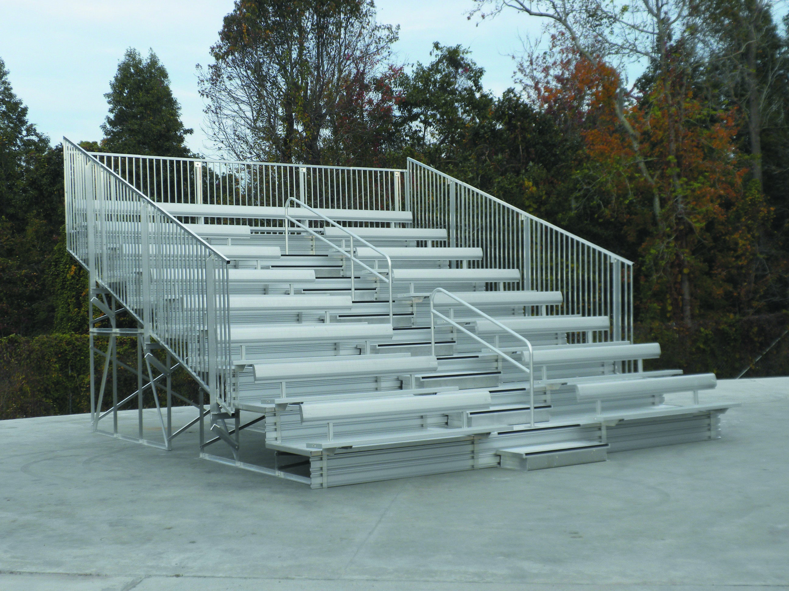 Eight-row galvanized steel bleachers with vertical picket guardrails and handrails on concrete surface surrounded by trees