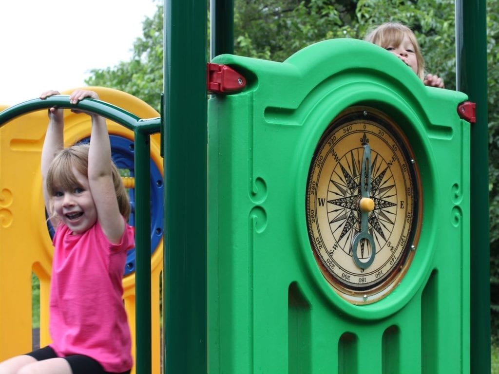 Green playground panel with large compass design, two children playing on colorful climbing structures outdoors