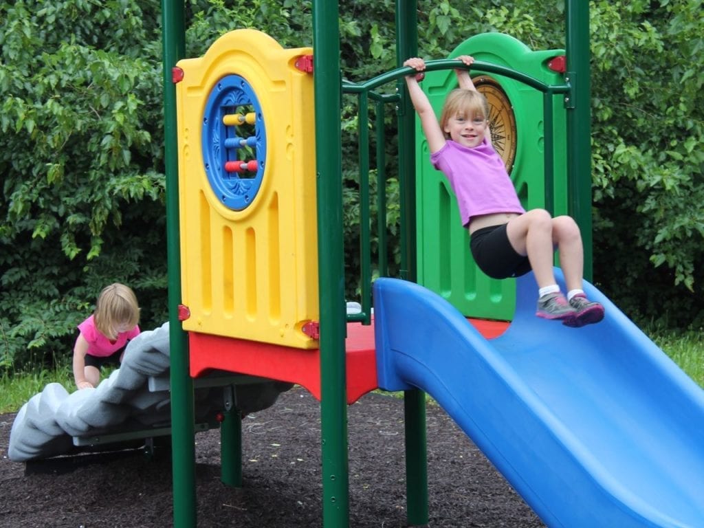 Colorful early childhood playground with blue river rapids slide, dinosaur fossil climber, activity panels, and roof structure.