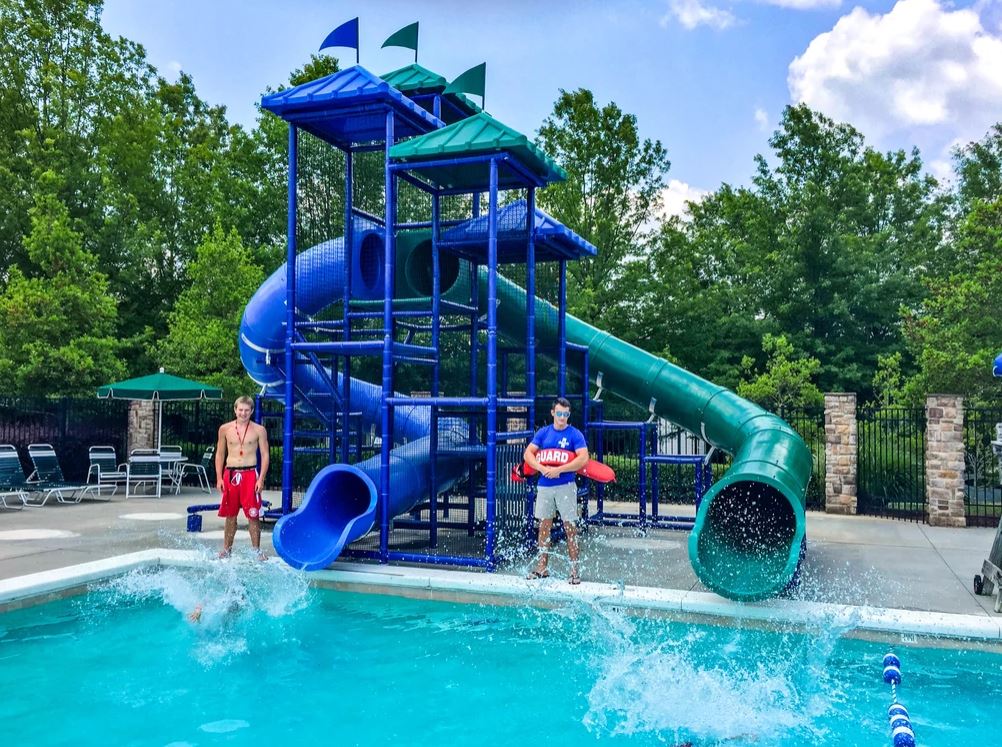 Two curved blue and green water slides with protective netting and roofs beside a pool with lifeguards and splash figures