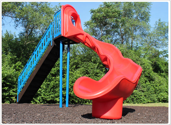 Red independent playground slide with blue stairs and curved design on mulch surface surrounded by green trees