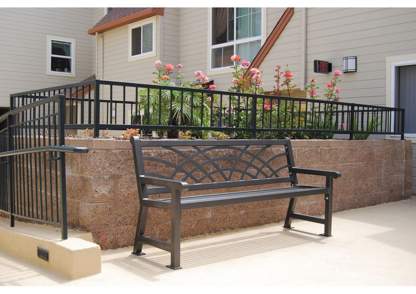 Outdoor metal bench with curved decorative backrest and armrests, placed on concrete near a raised flower garden bed