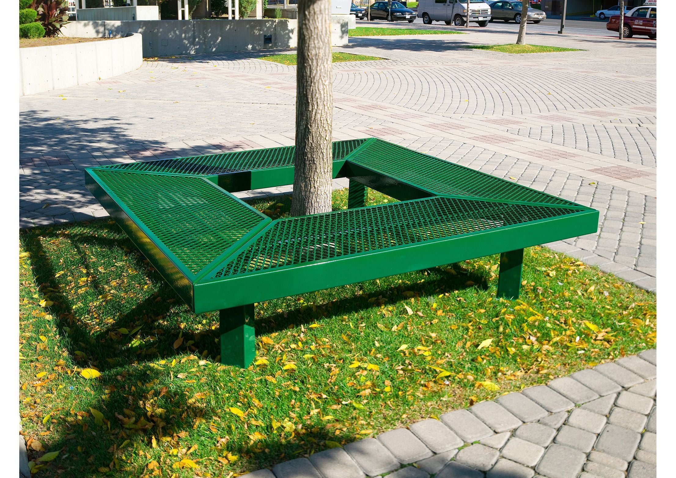 Green powder-coated metal square bench without backrest surrounding a tree on grass and paved outdoor area