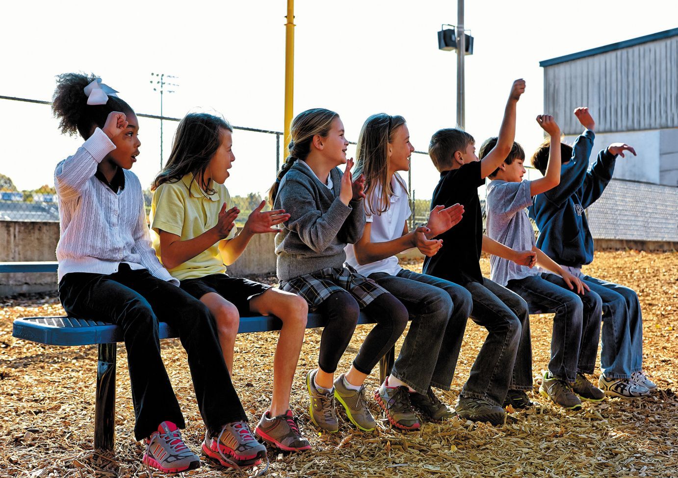 Heavy-duty perforated metal bench without backrest seating seven children on wood chip ground at sports field