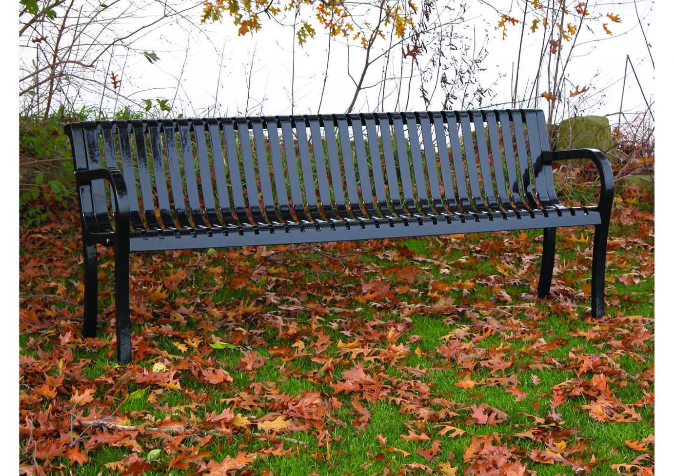 Black metal outdoor bench with backrest and armrests on grass with scattered autumn leaves