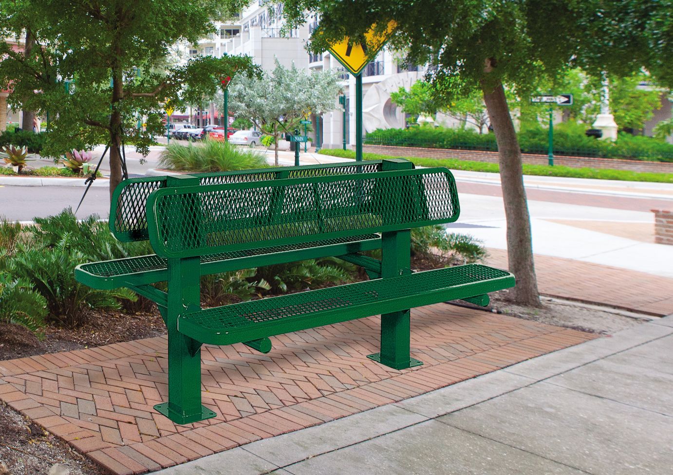 Green double-sided metal park bench with diamond mesh design, sturdy square posts, and backrests on brick-paved sidewalk