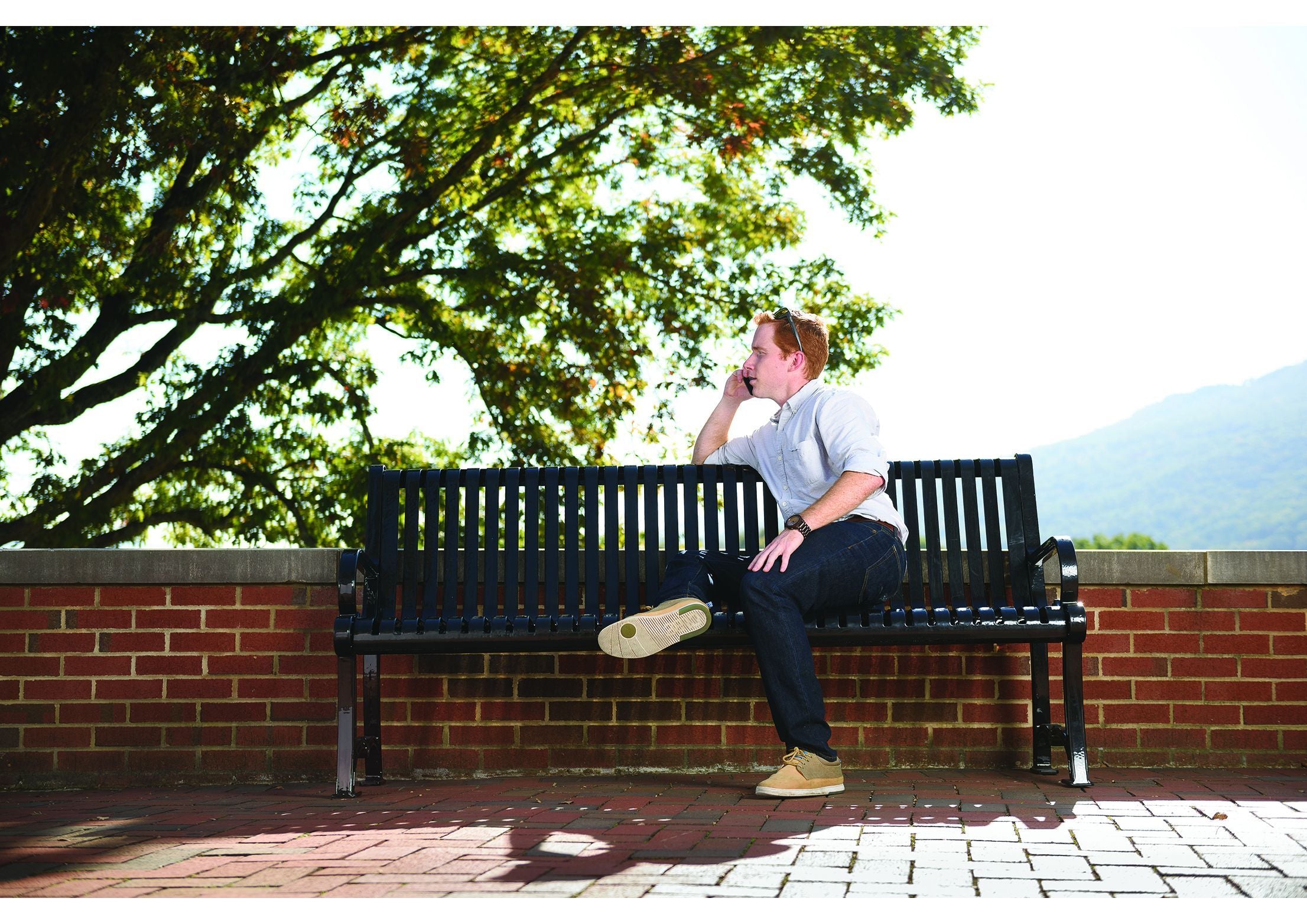 Black metal outdoor bench with ornate aluminum frame and backrest on brick patio, man seated and talking on phone, leafy tree and hills nearby